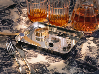 A styled tabletop scene with a silver tray holding several vintage diamond wristwatches and bracelets, alongside cutlery and two glasses of whiskey, all arranged on a black-and-white patterned cloth.