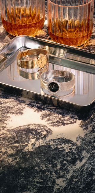 Two luxury wristwatches, one gold and one silver, displayed on a reflective silver tray with whiskey glasses in the background.