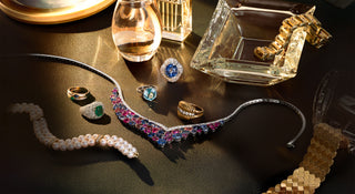 A luxury jewelry flat-lay featuring a prominent platinum sapphire necklace embellished with pink, blue, and purple sapphires, alongside gold rings, a pearl strand, and other vintage and antique accessories on a dark reflective surface.