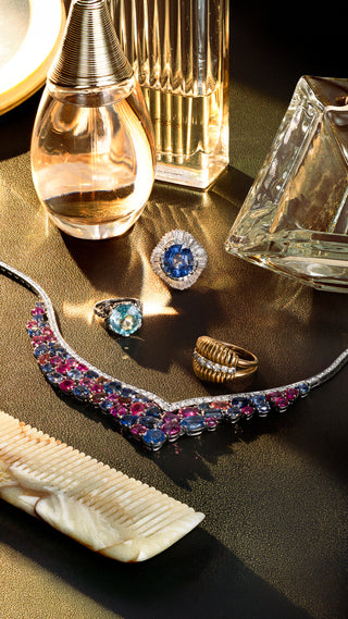 A luxury jewelry flat-lay featuring a silver-toned collar necklace adorned with pink and blue gemstones, alongside gold rings, a blue gemstone ring, and a white shell comb, styled with perfume bottles on a dark reflective surface.