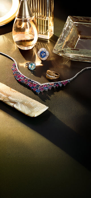 A styled flat-lay of vintage jewelry on a dark reflective surface, featuring a prominent platinum sapphire necklace embellished with pink and blue sapphires, alongside rings and other accessories.