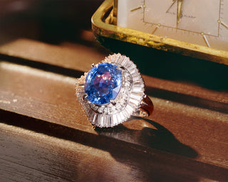 A luxury platinum blue sapphire ring featuring a large oval blue sapphire center stone surrounded by a halo of baguette-cut diamonds and smaller round diamonds, set on a polished band. The ring is displayed on a dark wooden surface next to a vintage gold pocket watch.