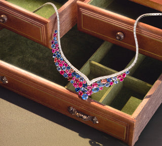 A platinum necklace adorned with vibrant pink, blue, and orange sapphires, displayed inside an open wooden jewelry box with green velvet compartments.