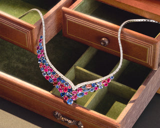 A V-shaped diamond necklace embellished with pink, blue, and red sapphires, displayed inside an open wooden jewelry box with green velvet compartments.