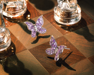 A pair of butterfly-shaped stud earrings set with purple sapphires on a polished wooden board, with glass chess pieces partially visible in the background.
