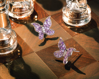 A pair of butterfly-shaped stud earrings paved with purple sapphires and set in white gold, displayed on a polished wooden chessboard with glass chess pieces.